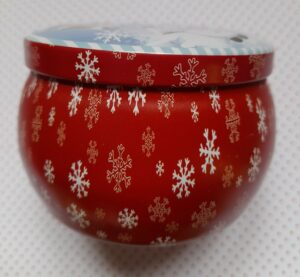 Vue de côté d'une boite ronde vintage contenant une bougie parfumée, la couleur est rouge et elle est décorée de divers flocons de neige de couleur blanche et de tailles différentes