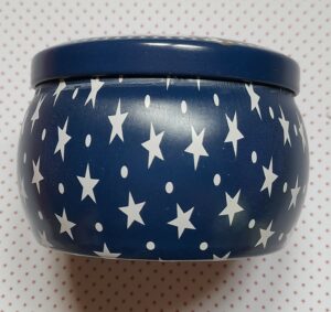 Vue de côté d'une boite ronde vintage contenant une bougie parfumée, la couleur est bleue nuit et elle est décorée d'étoiles et de points blanc