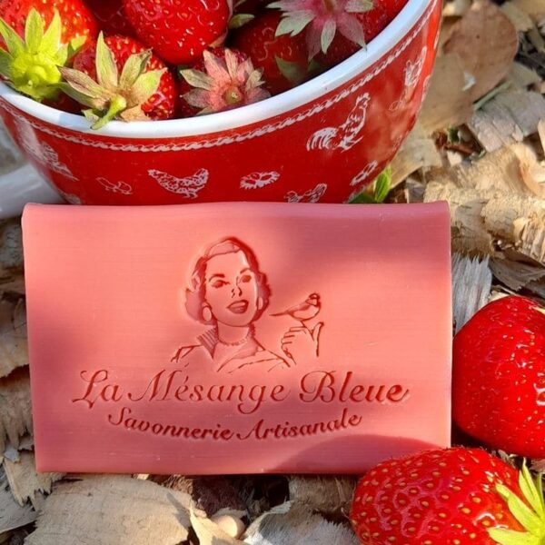 Savon artisanal naturel parfumé à la fraise, posé au milieu de fraisiers sur des copeaux de bois devant un bol de fraises bien rouges.