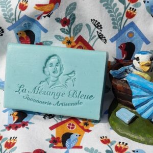 Savon artisanal naturel au nom de "La Mésange Bleue" dont le parfum est secret. Ce savon est posé sur un essuie-mains avec des motifs de nids et d'oiseaux bleus, à côté du savon se trouve une mésange en terre cuite qui lave son petit dans une baignoire médiévale.