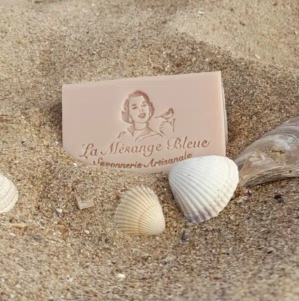 Savon artisanal naturel parfumé au monoï posé dans le sable sur la plage avec des coquillages devant le savon.