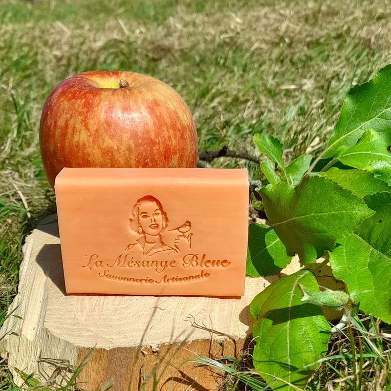 Savon artisanal naturel parfumé à la pomme, posé sur une tranche de bois brut devant une pomme fruit à coté du branche de pommier. Dans un champ en herbe, un jour de soleil.