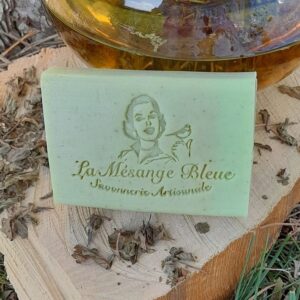 Savon artisanal naturel parfumé à la verveine posé sur un rondin de bois adossé à une théière emplie d'infusion de verveine et des feuilles de verveine séchées tout autour du savon.