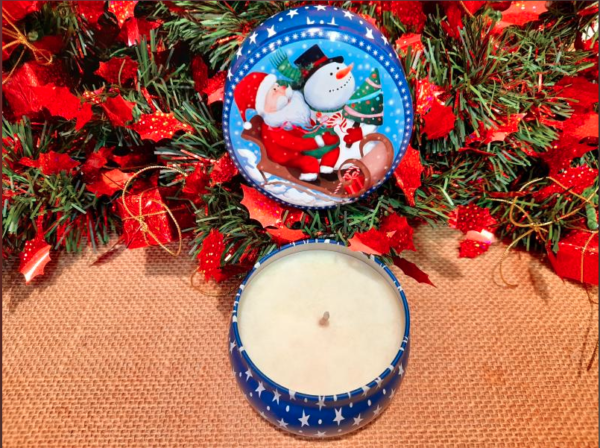 Photo d'une bougie parfumée dans une boîte ronde vintage avec en décor un père noël, un bonhomme de neige et un sapin sur un traineau, le tout dans un décor de noël