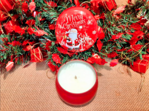 Photo d'une bougie parfumée dans une boîte ronde rouge vintage ouverte avec en décor une illustration d'un père noël, de flocons de neiges, de cadeaux et d'une inscription "merry christmas", le tout dans un décor de noël