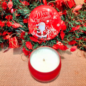 Photo d'une bougie parfumée dans une boîte ronde rouge vintage ouverte avec en décor une illustration d'un père noël, de flocons de neiges, de cadeaux et d'une inscription "merry christmas", le tout dans un décor de noël
