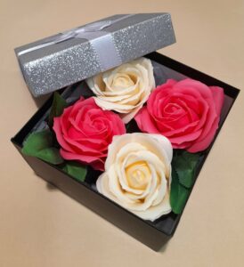 Vue intérieure d'une boîte cadeau noire avec le couvercle gris pailleté et un ruban argenté autour, contenant 4 savons en forme de roses, 2 de couleur blanche et 2 de couleur rose