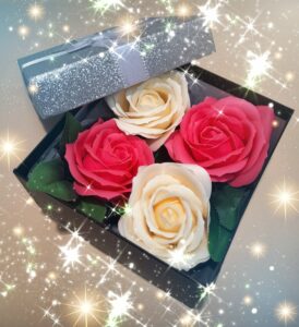 Boîte cadeau noire avec le couvercle gris pailleté et un ruban argenté autour, contenant 4 savons en forme de roses, 2 de couleur blanche et 2 de couleur rose
