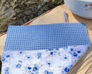 Essuie-mains modèle Jacques, aux motifs floraux dans les tons bleus dont on voit le verso bleu qui est en matière nid d'abeille, posé sur un plan de travail en bois avec une vasque vintage, dans un décor naturel