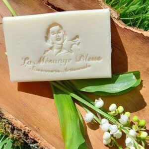 Savon artisanal naturel parfumé au muguet, posé dans un morceau d'écorce de bois brut, avec un brin de muguet. Décor champêtre ensoleillé.