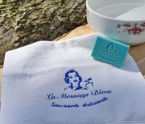 Serviette blanche brodée au logo de la Savonnerie "La Mésange Bleue", posée sur un plan de travail en bois à côté d'une vasque vintage avec un savon bleu turquoise dans un décor naturel.