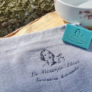 Serviette brodée grise à l'effigie de la savonnerie la mésange bleue, posée sur une planche en bois avec une vasque vintage et un savon turquoise dans un cadre extérieur bucolique