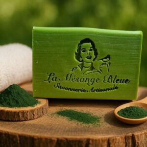 Savon artisanal naturel à la spiruline BIO de la Savonnerie La Mésange Bleue. Posé sur une tranche de bois avec de la poudre de spiruline dans un décor naturel de verdure coordonné à la couleur verte du savon.